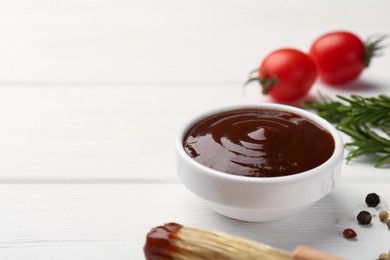 Photo of Tasty barbeque sauce in bowl, rosemary and spices on white wooden table, closeup. Space for text