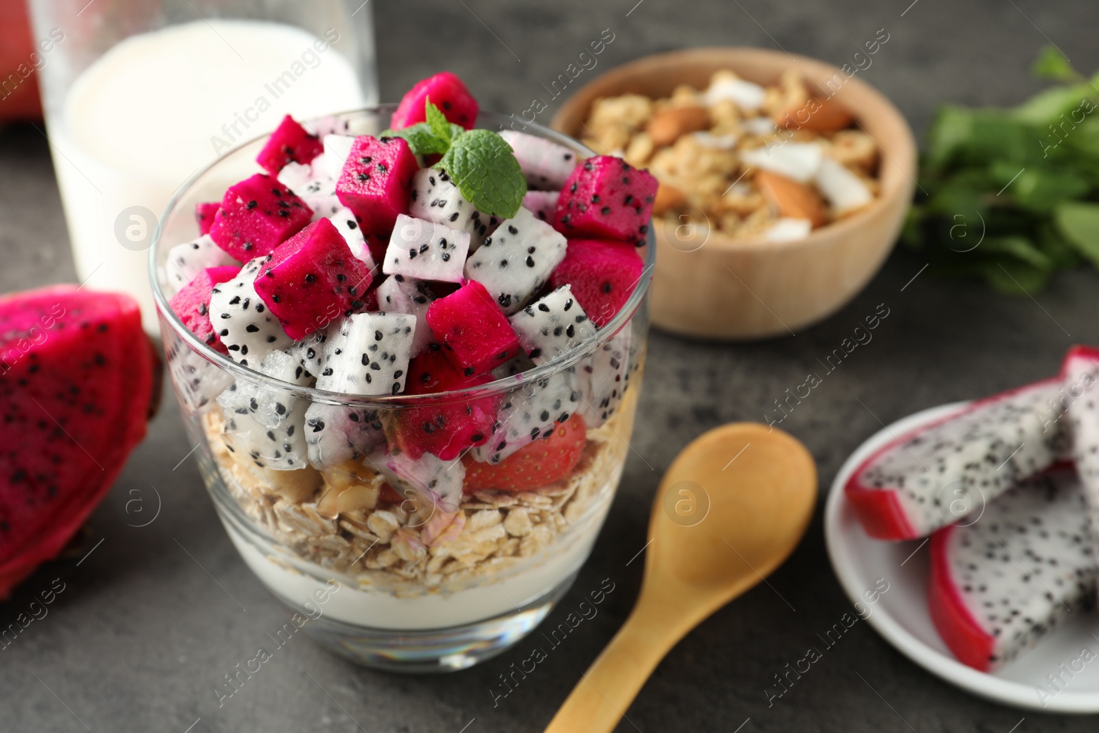 Photo of Glass of granola with different pitahayas, yogurt and other fruits on grey table, space for text