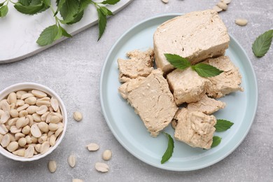Photo of Plate with pieces of tasty halva, peanuts and mint leaves on light grey table, flat lay