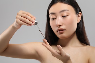 Photo of Beautiful young woman applying cosmetic serum onto her finger on grey background, selective focus
