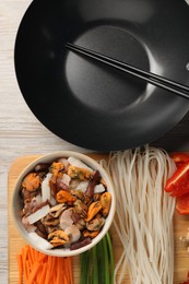 Photo of Black wok, chopsticks and board with products on light wooden table, top view