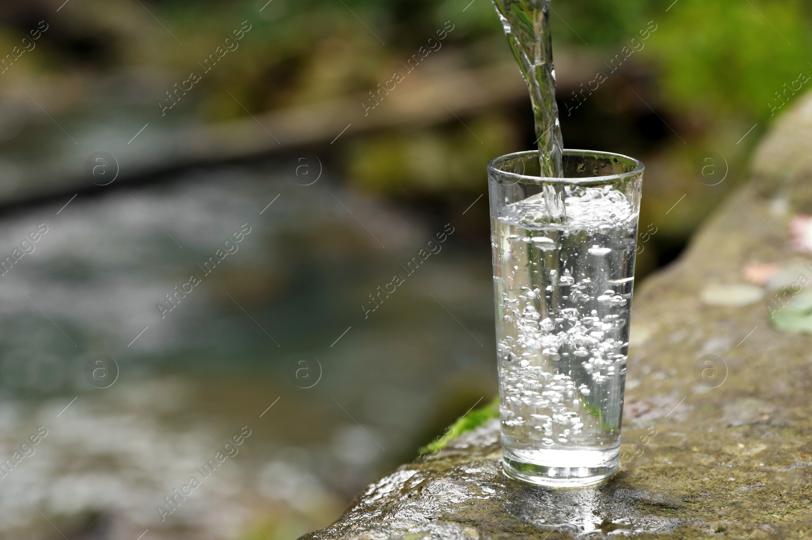 Photo of Fresh water pouring into glass on stone near river. Space for text