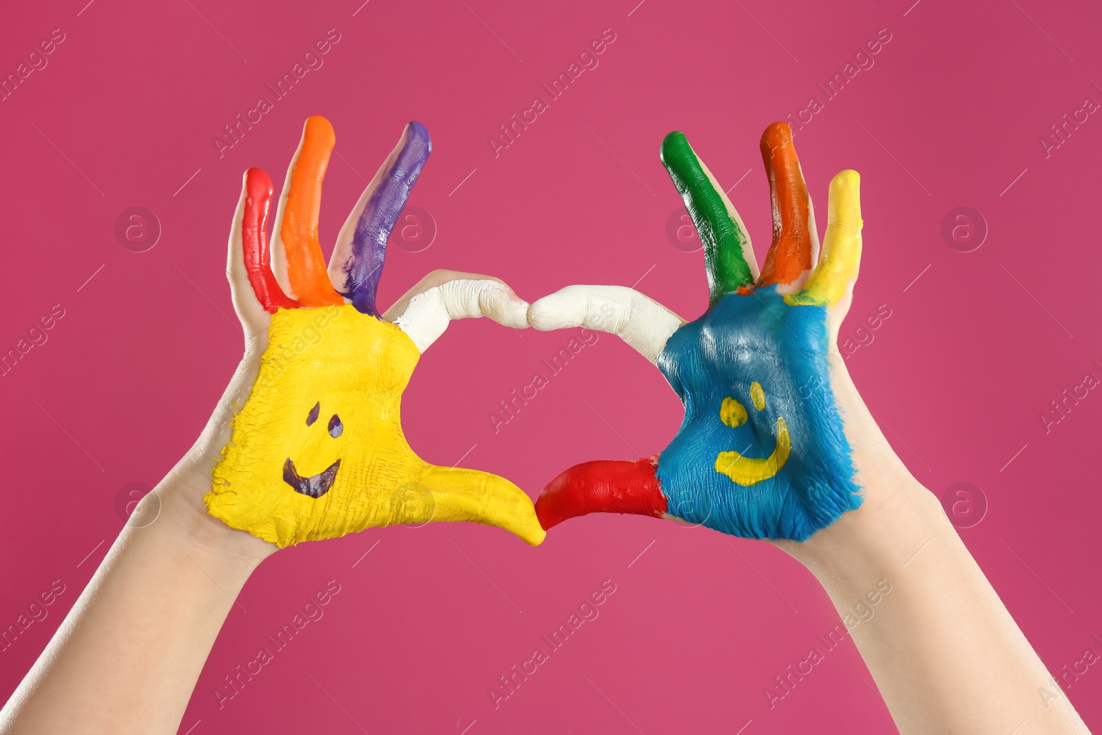 Photo of Kid with smiling faces drawn on palms showing heart gesture against pink background, closeup