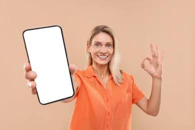 Happy woman holding smartphone with blank screen and showing OK gesture on beige background