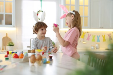 Photo of Easter celebration. Cute children with bunny ears painting eggs at white table in kitchen