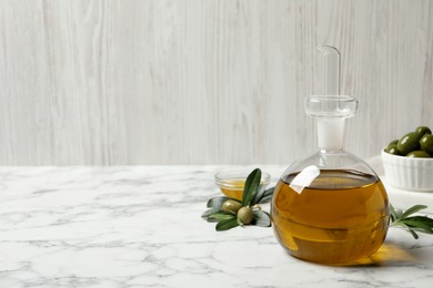 Photo of Glass jug of oil, ripe olives and green leaves on white marble table. Space for text