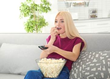 Photo of Young woman with remote control and bowl of popcorn watching TV on sofa at home