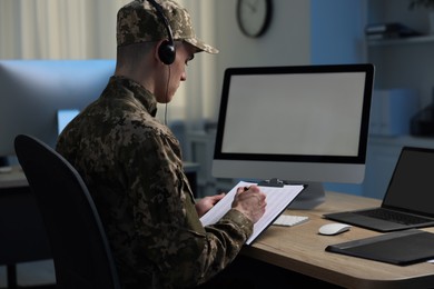 Photo of Military service. Soldier with clipboard and headphones working at table in office at night