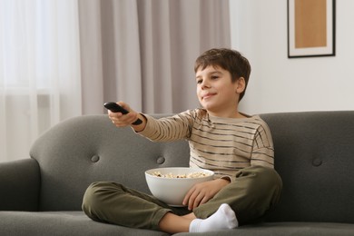 Little boy holding bowl of popcorn and changing TV channels with remote control on sofa at home