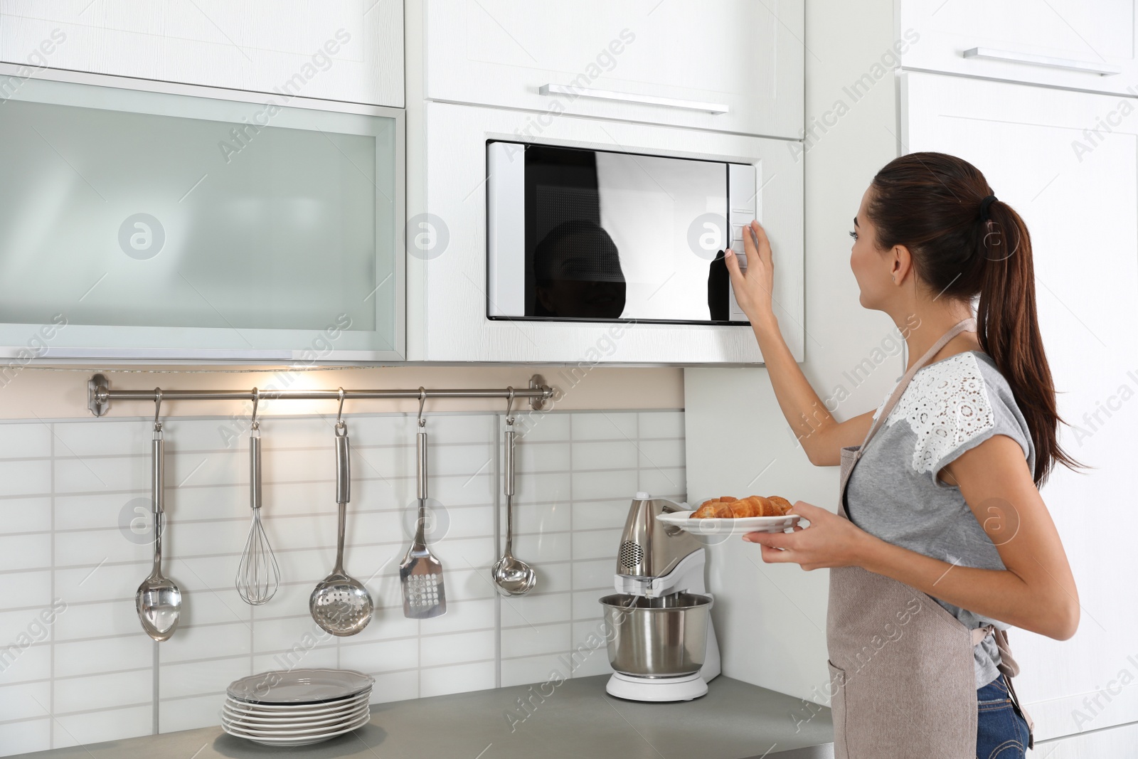 Photo of Young woman with plate of croissants near microwave oven in kitchen