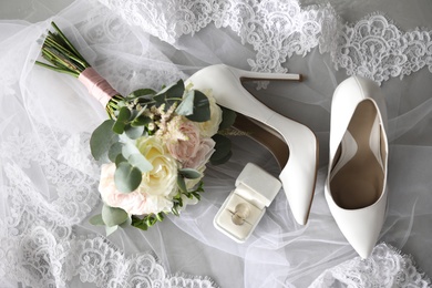 Photo of Pair of white high heel shoes, veil, rings and wedding bouquet on grey background, flat lay