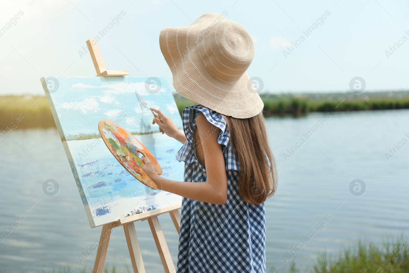 Photo of Little girl painting scenery on easel near lake, back view