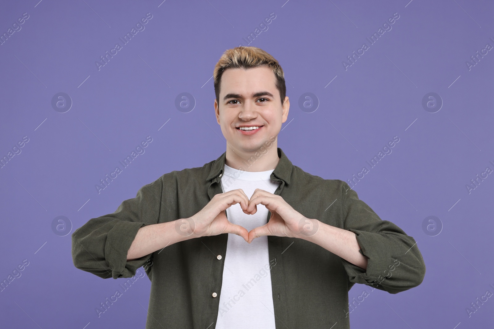 Photo of Young man showing heart gesture with hands on purple background
