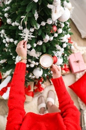 Woman with cup of delicious hot drink decorating Christmas tree at home, top view