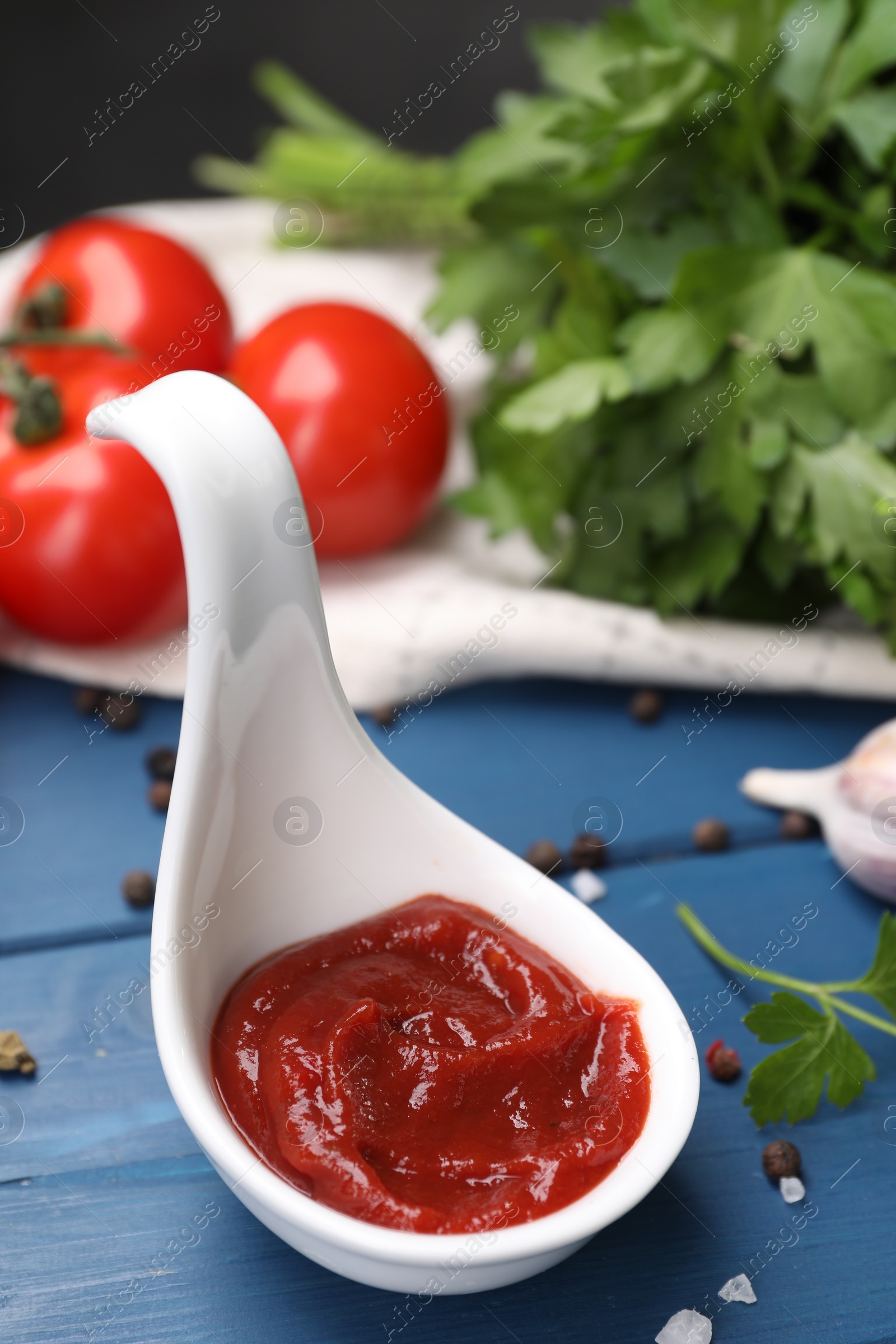 Photo of Organic ketchup in spoon and spices on blue wooden table, closeup. Tomato sauce