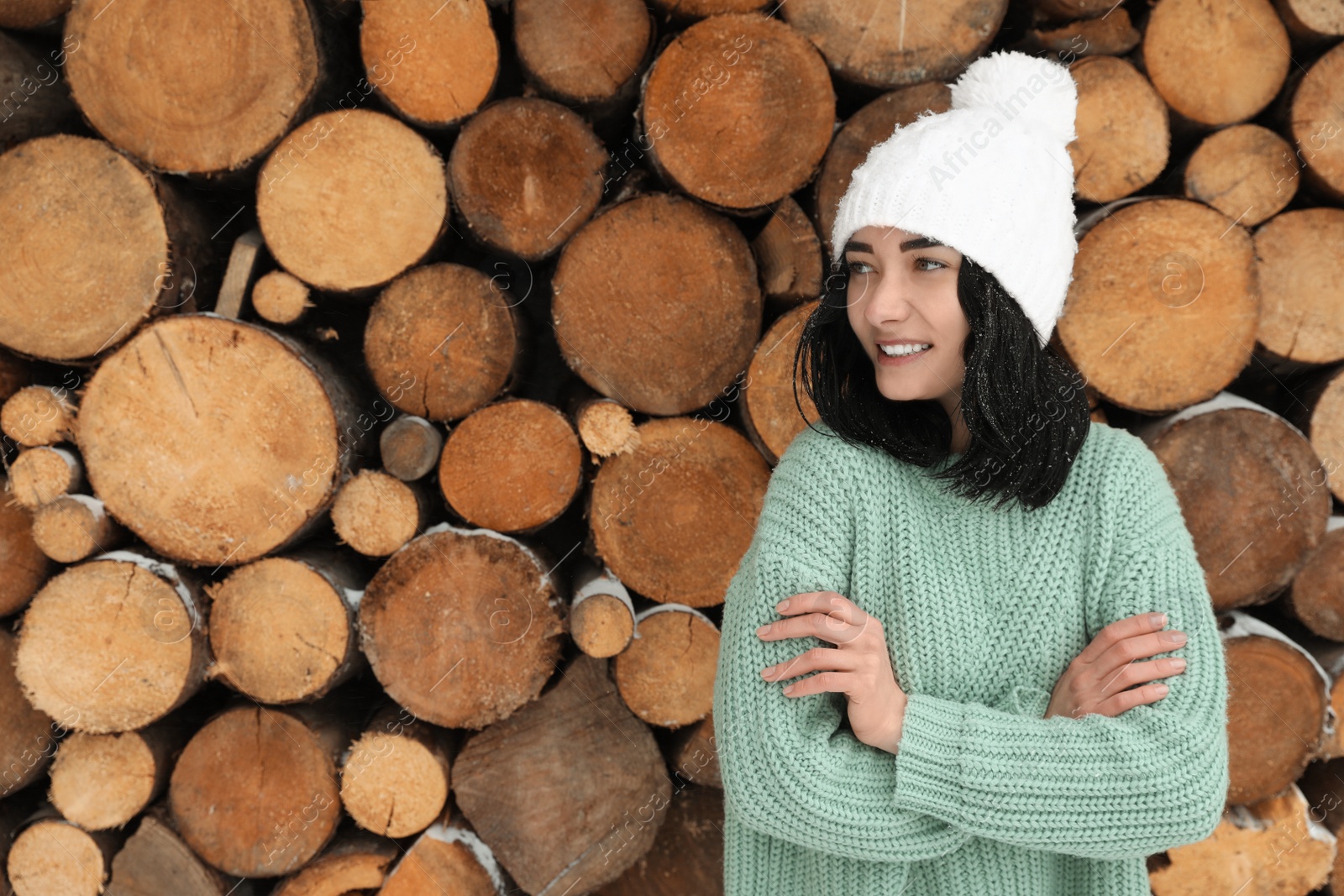 Photo of Young woman wearing warm sweater and hat near stack of firewood, space for text. Winter season