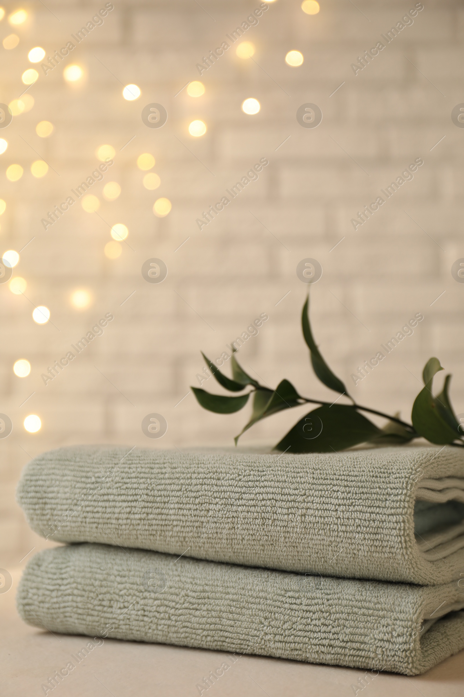 Photo of Stacked soft towels and green branch on white table near brick wall indoors, space for text