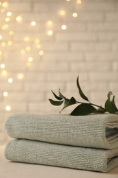 Photo of Stacked soft towels and green branch on white table near brick wall indoors, space for text