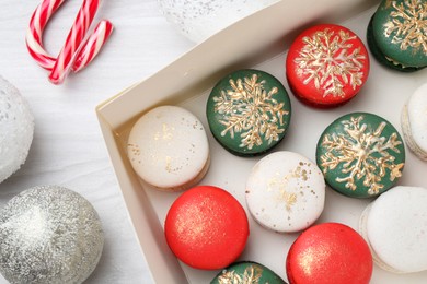 Photo of Different decorated Christmas macarons in box and festive decor on white wooden table, flat lay