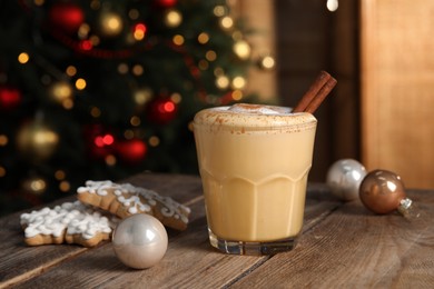 Photo of Tasty eggnog with cinnamon, cookies and Christmas baubles on wooden table against blurred festive lights. Space for text