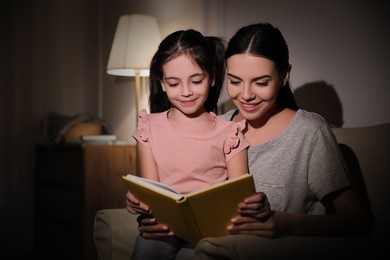 Photo of Little girl with mother reading fairy tale at home in evening