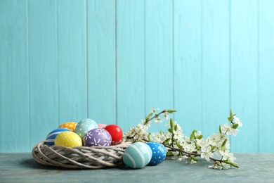 Photo of Painted Easter eggs in nest and blossoming branches on table against color background. Space for text