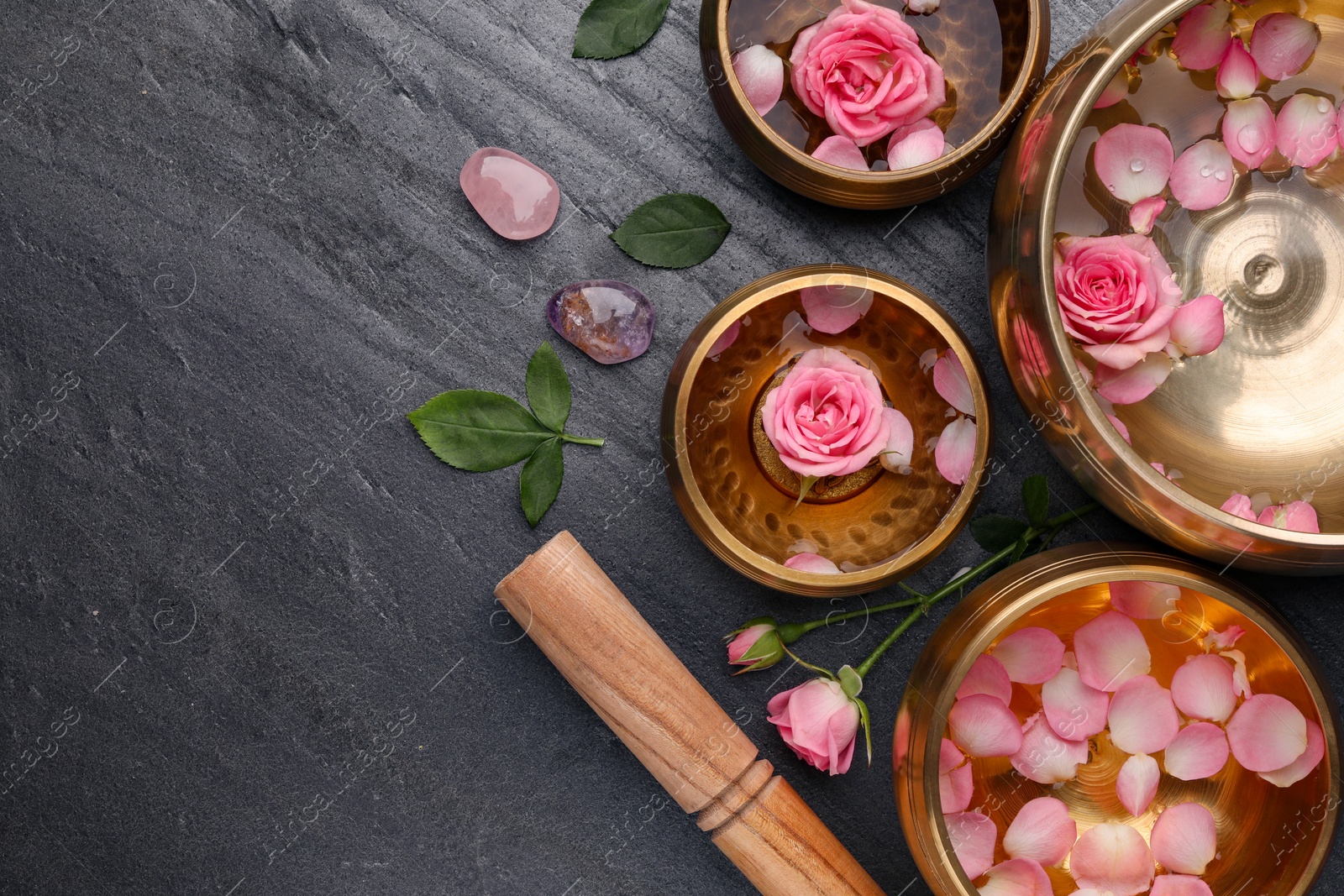 Photo of Tibetan singing bowls with water, beautiful rose flowers and mallet on gray table, flat lay. Space for text