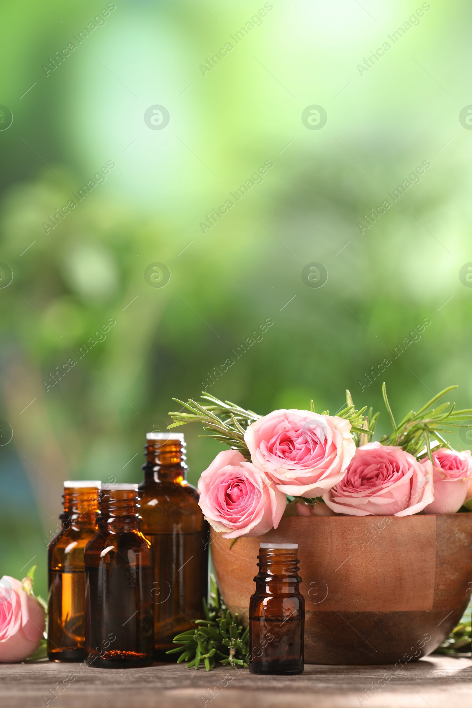 Photo of Bottles with essential oils, roses and rosemary on wooden table against blurred green background. Space for text