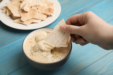 Photo of Woman dipping pita chip into hummus at turquoise wooden table, closeup