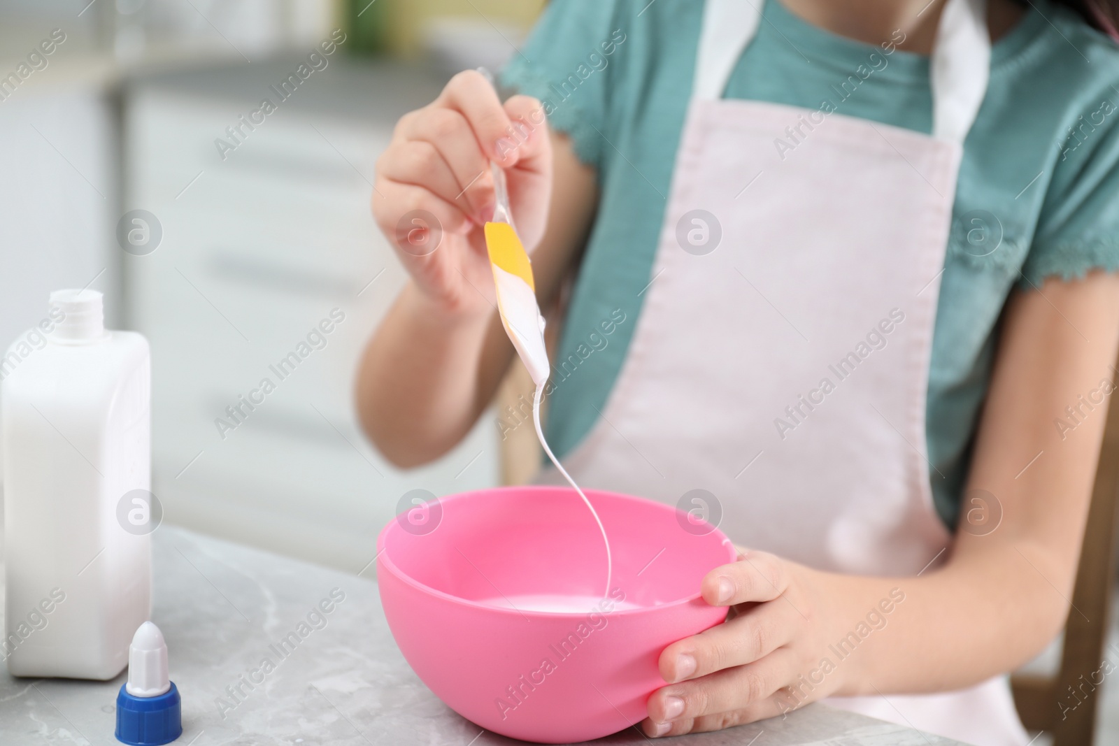 Photo of Little girl mixing ingredients with silicone spatula at table in kitchen, closeup. DIY slime toy