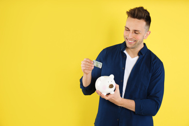Happy man putting money into piggybank on yellow background. Space for text