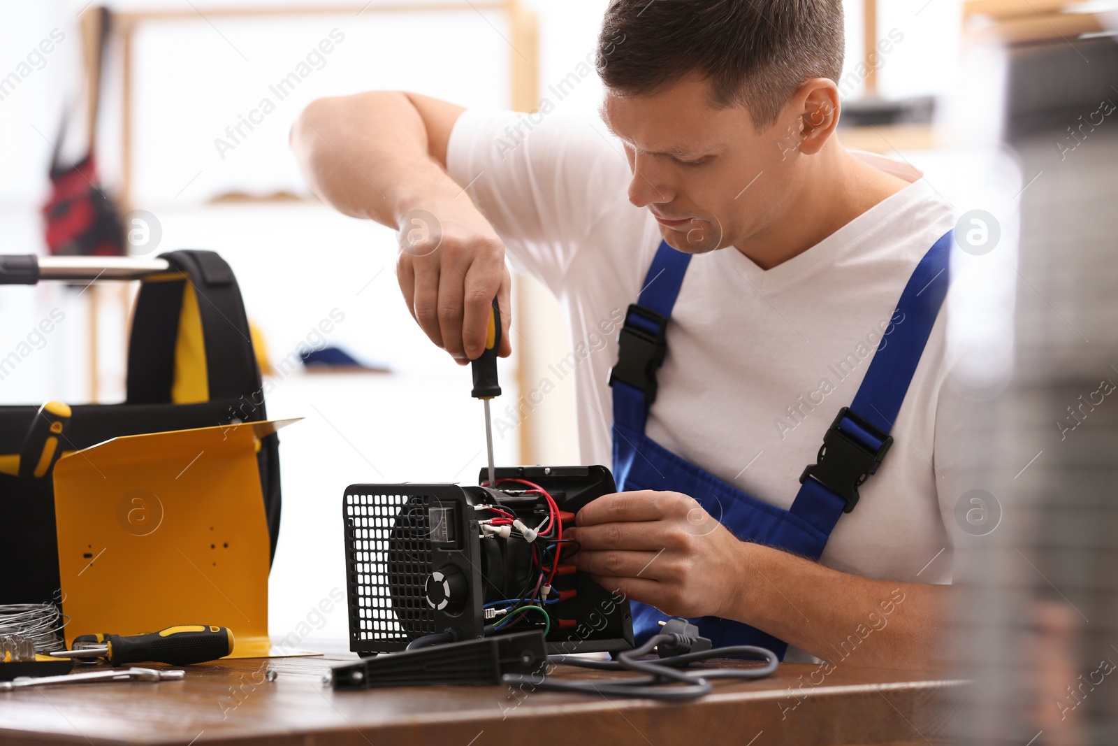 Photo of Professional technician repairing electric fan heater with screwdriver at table indoors