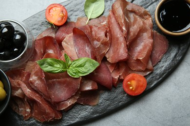 Photo of Delicious bresaola, tomato, olives and basil leaves on grey textured table, top view