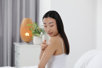 Photo of Beautiful young Asian woman applying body cream on shoulder in bedroom