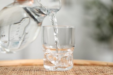 Pouring fresh water from jug into glass at wicker surface against blurred background, closeup