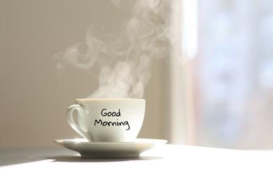 Image of Delicious coffee in cup with Good Morning wish on sunlit table. Space for text