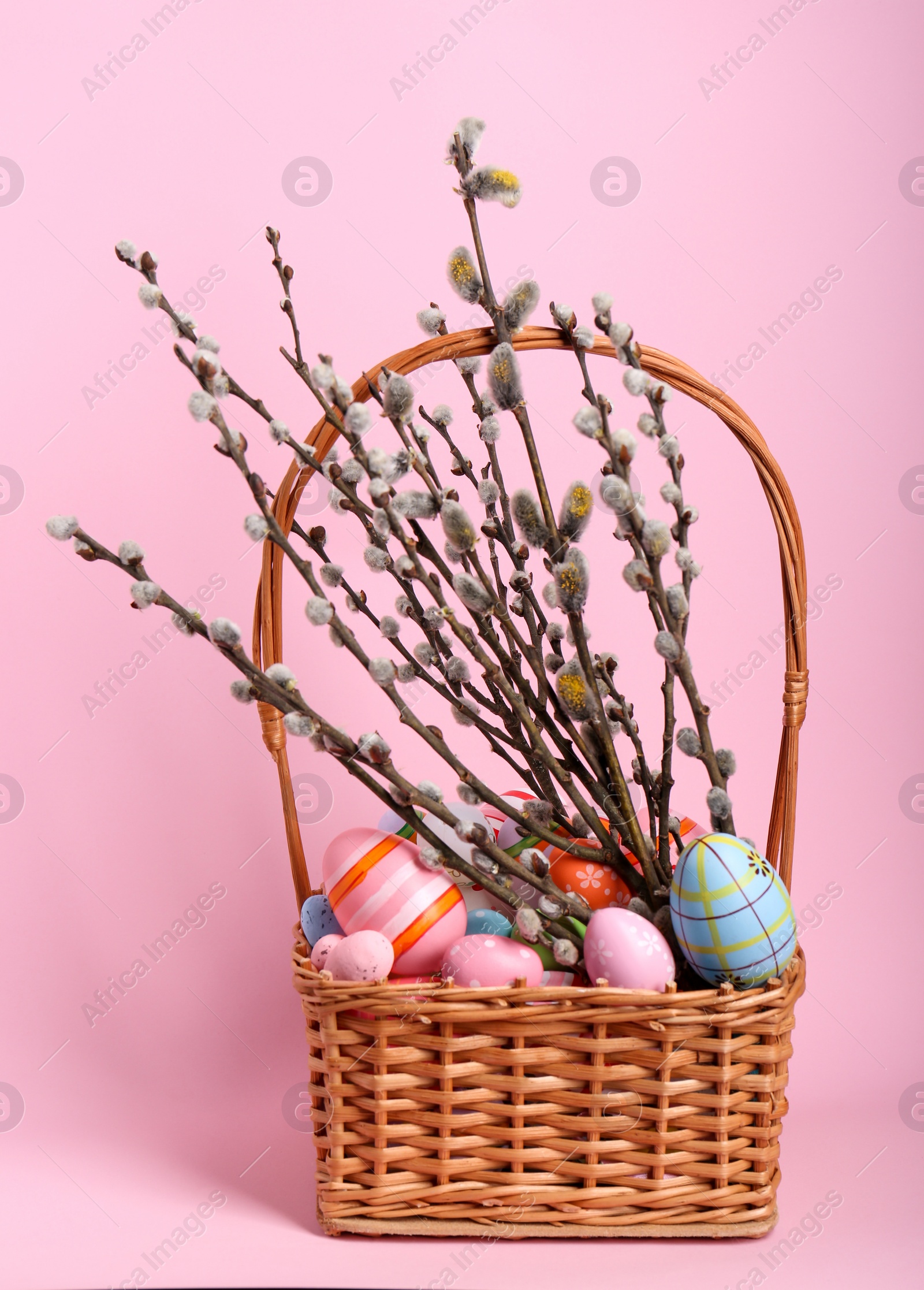 Photo of Wicker basket with beautiful willow branches and painted eggs on pink background. Easter decor