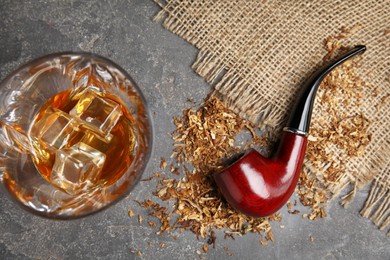 Photo of Smoking pipe with tobacco and glass of whiskey on grey table, flat lay