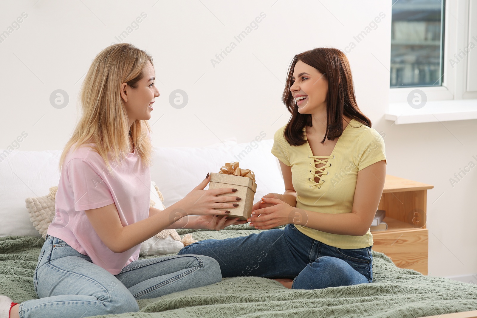 Photo of Smiling young woman presenting gift to her friend on bed at home