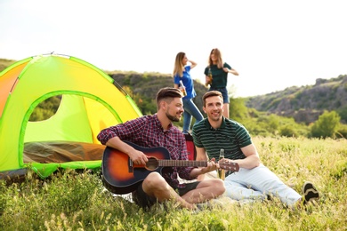 Group of young people resting with beer and guitar near camping tent in wilderness