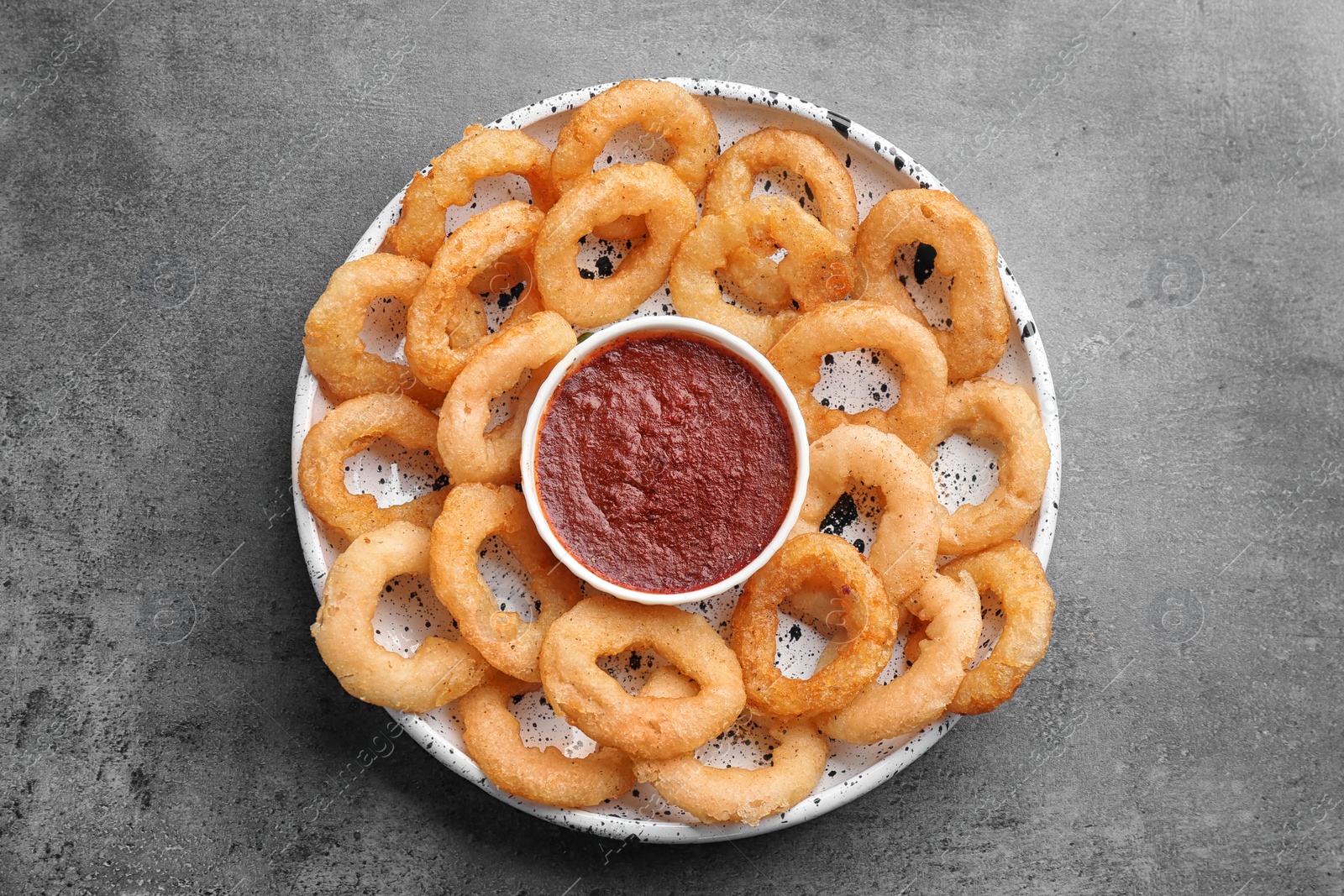 Photo of Plate with fried onion rings and bowl of sauce on grey background, top view