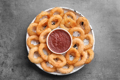 Plate with fried onion rings and bowl of sauce on grey background, top view