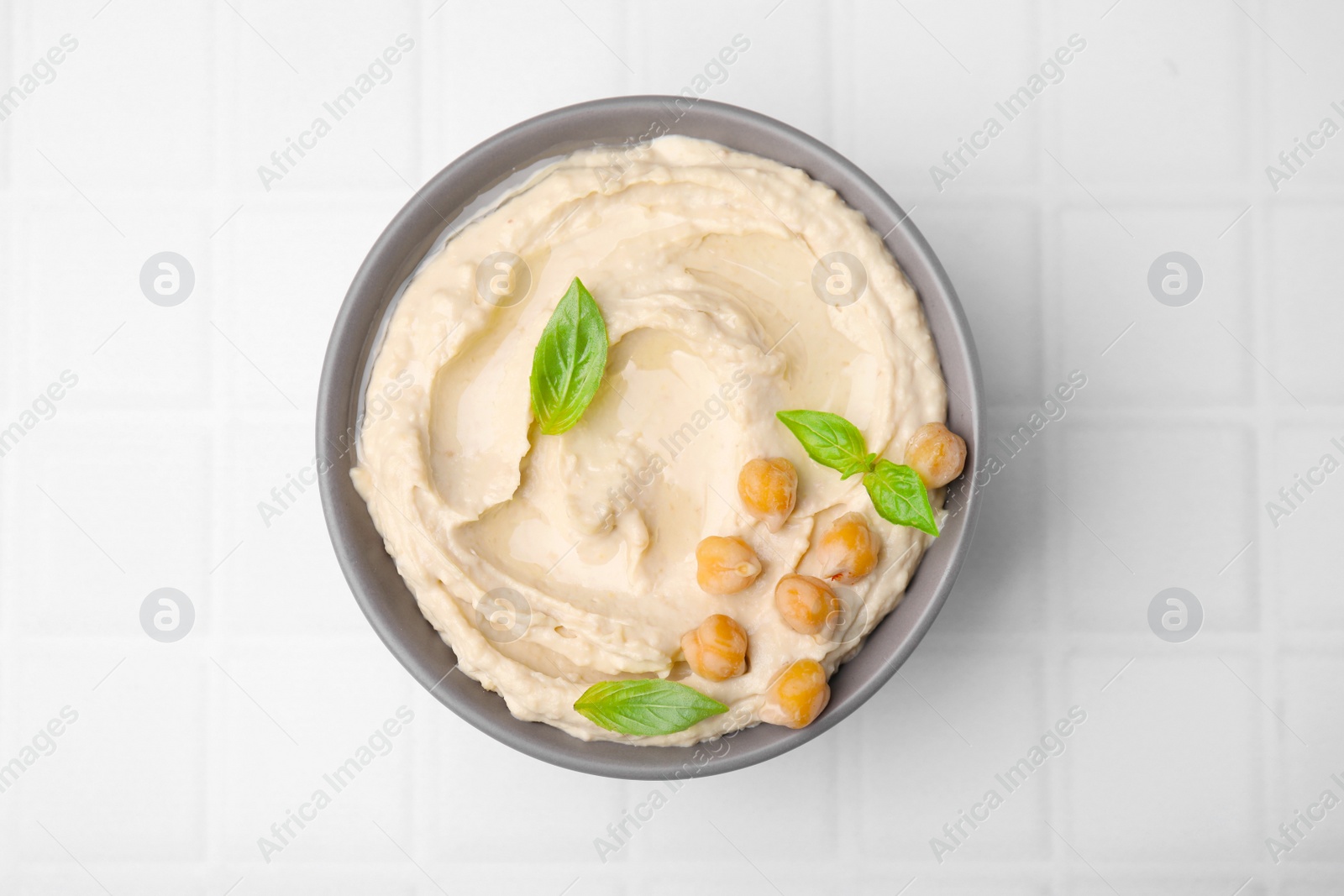 Photo of Bowl of delicious hummus with chickpeas on white tiled table, top view