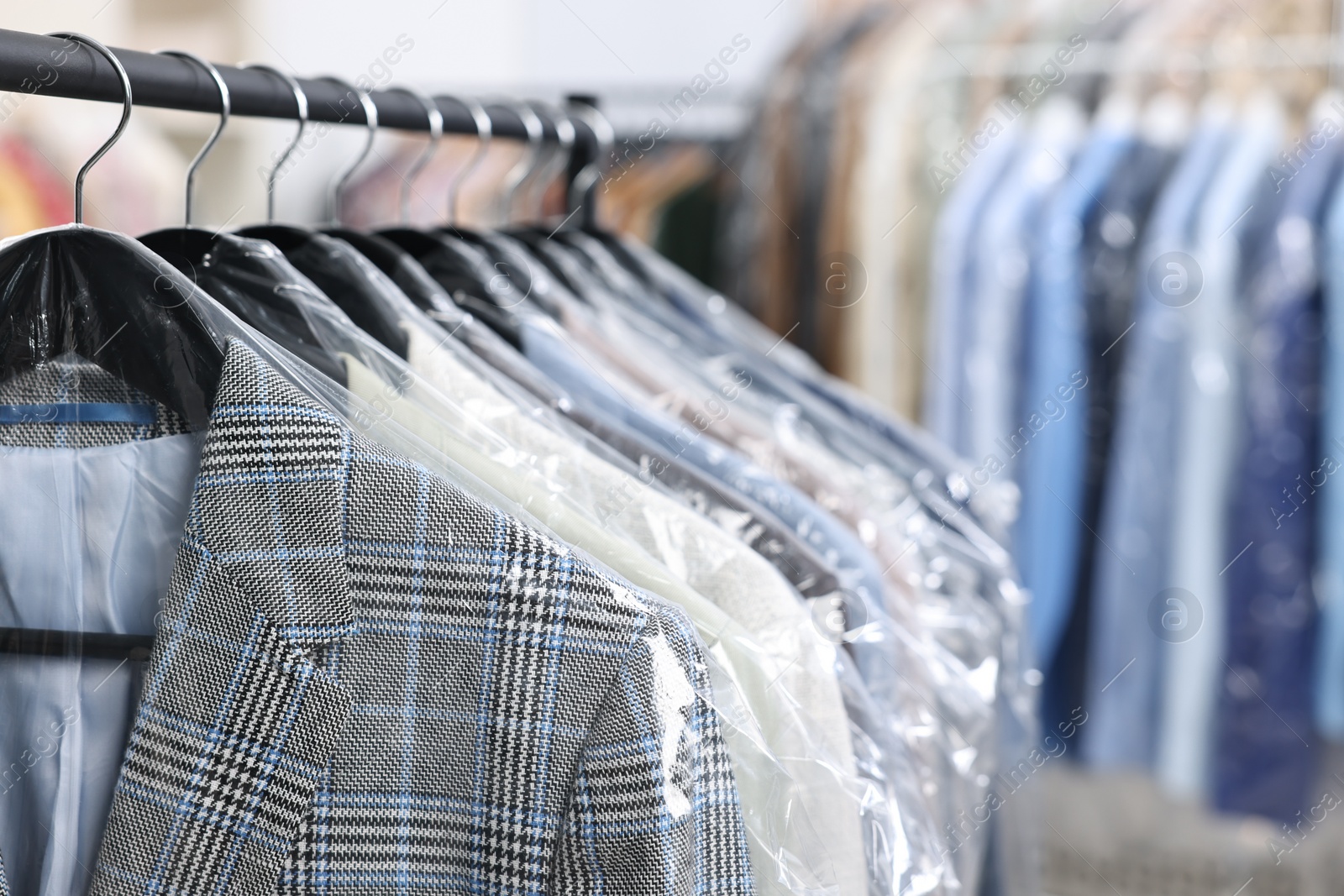 Photo of Dry-cleaning service. Many different clothes in plastic bags hanging on rack indoors, closeup