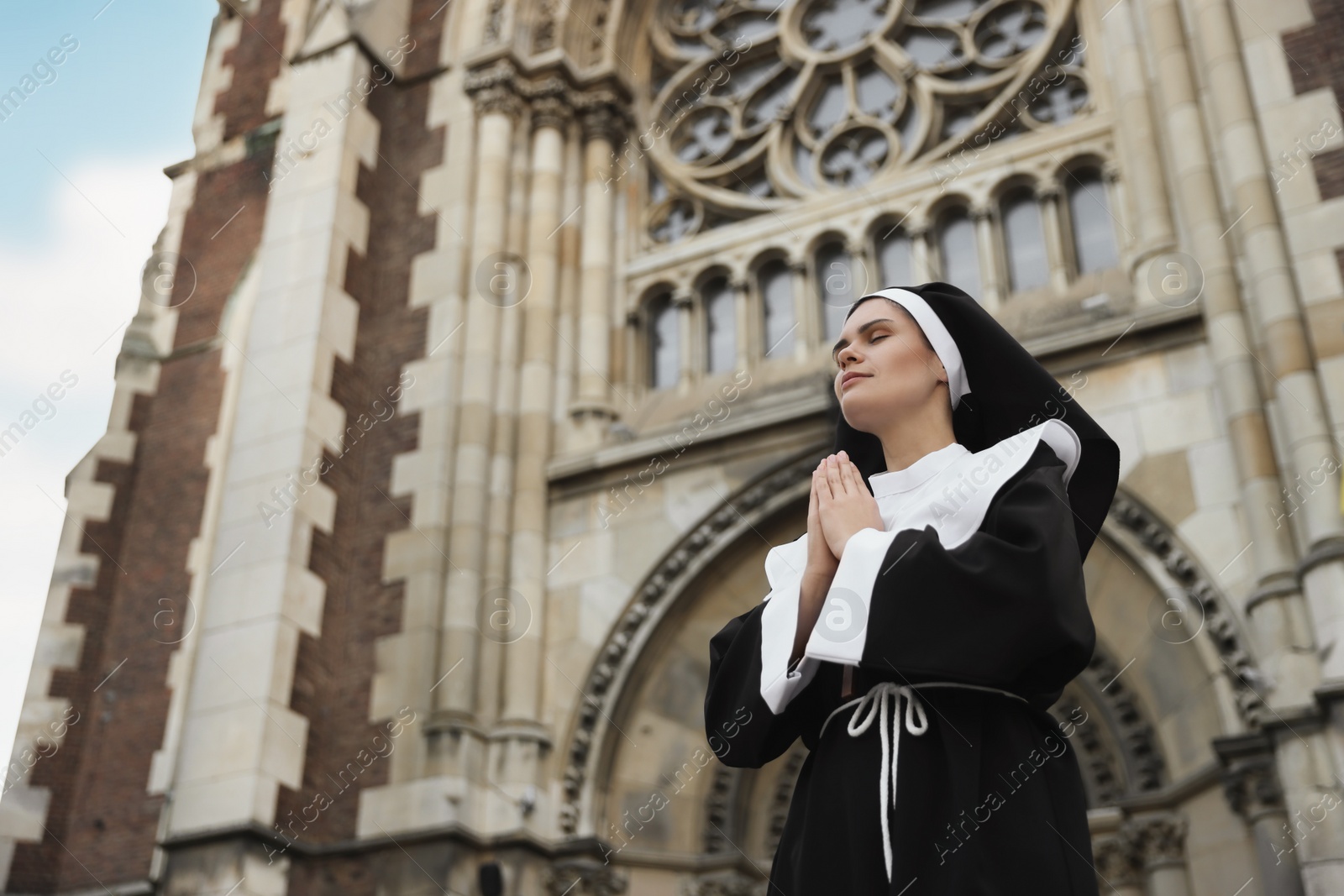 Photo of Young nun with hands clasped together while praying near cathedral outdoors, low angle view. Space for text
