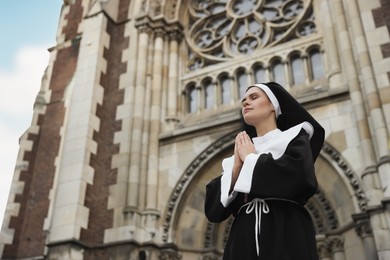 Photo of Young nun with hands clasped together while praying near cathedral outdoors, low angle view. Space for text