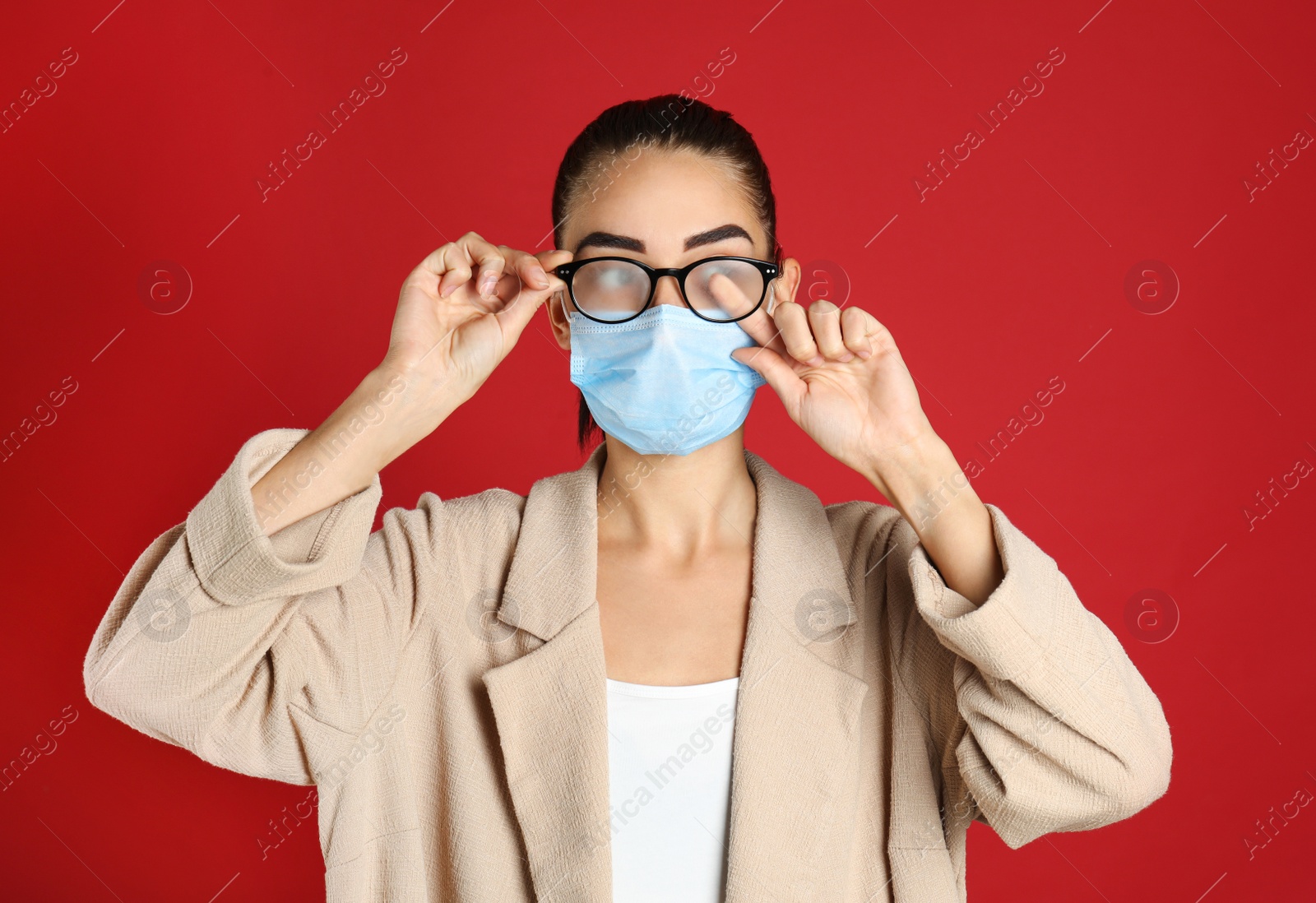 Photo of Woman wiping foggy glasses caused by wearing medical mask on red background