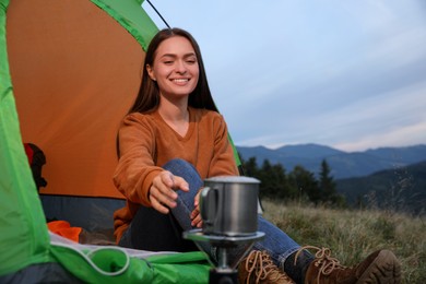 Photo of Young woman taking cup off stove while sitting in camping tent outdoors