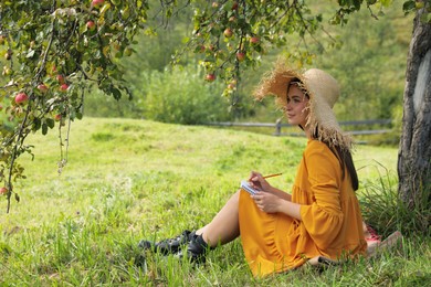 Photo of Beautiful young woman drawing with pencil in notepad on green grass near apple tree