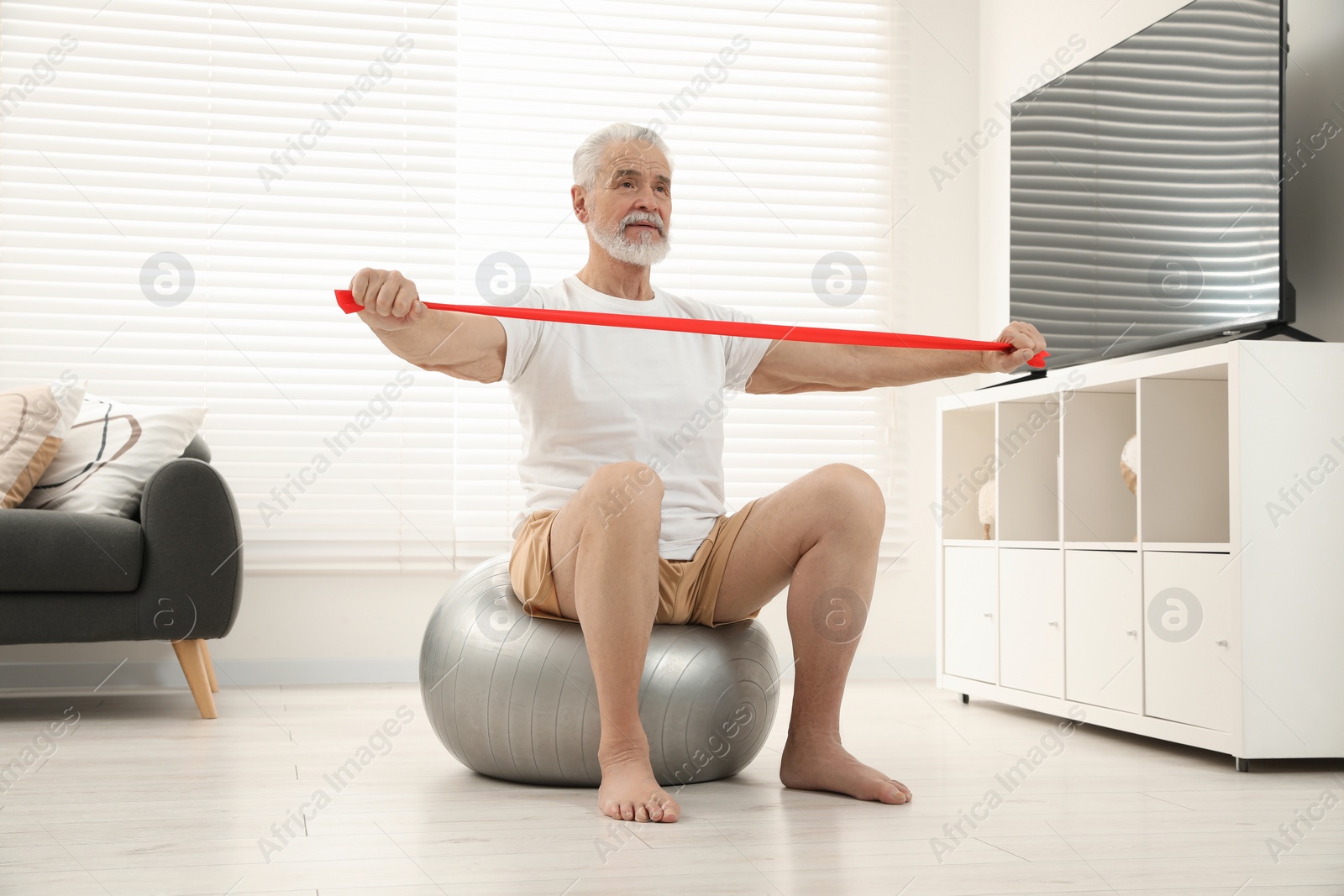 Photo of Senior man doing exercise with elastic resistance band on fitness ball at home